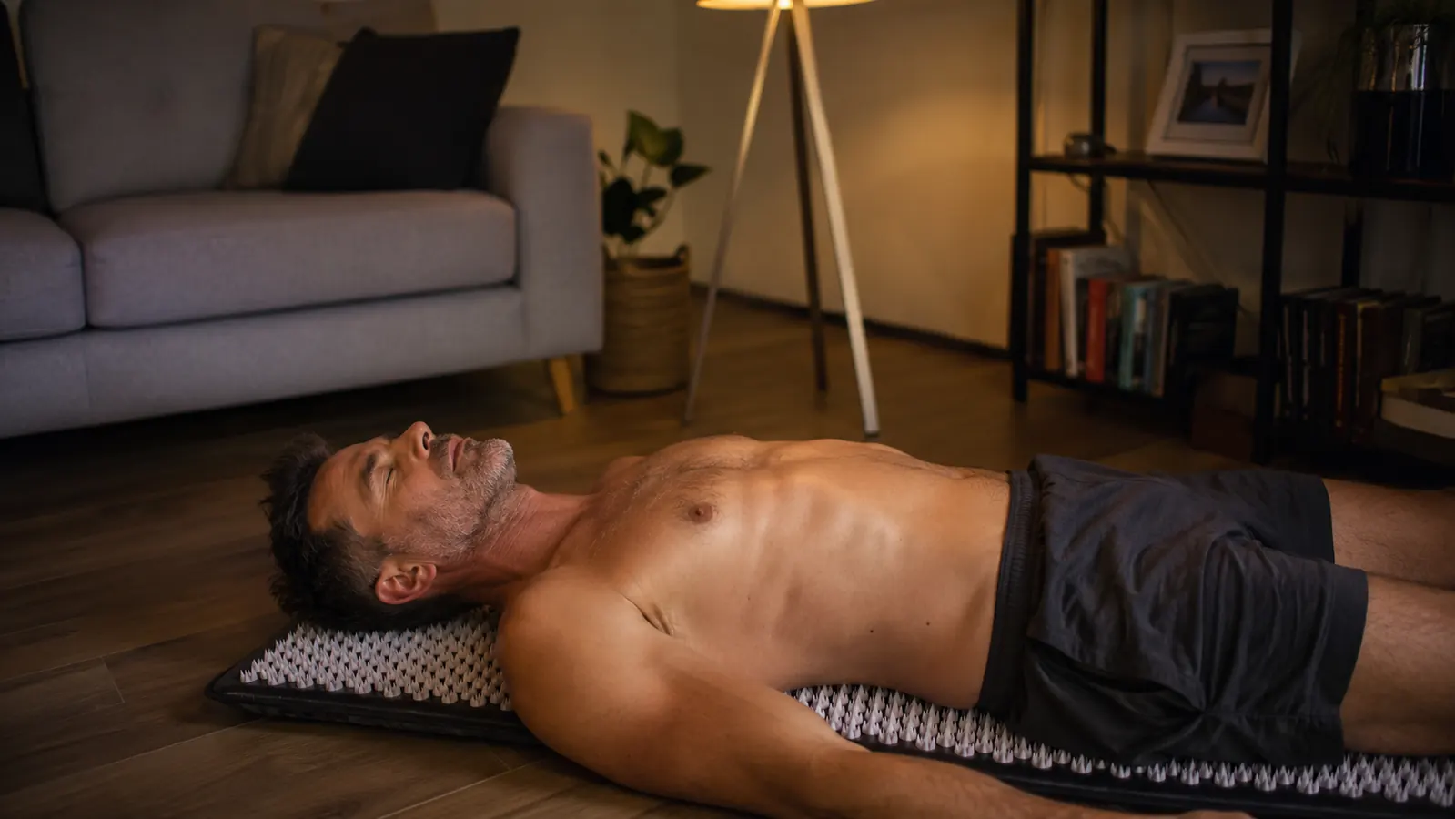 Man in his 40s lying on an acupressure mat on the living room floor with eyes closed, evening light through the window