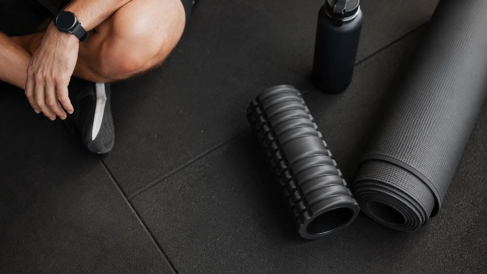 A foam roller resting on a gym floor next to a yoga mat and water bottle