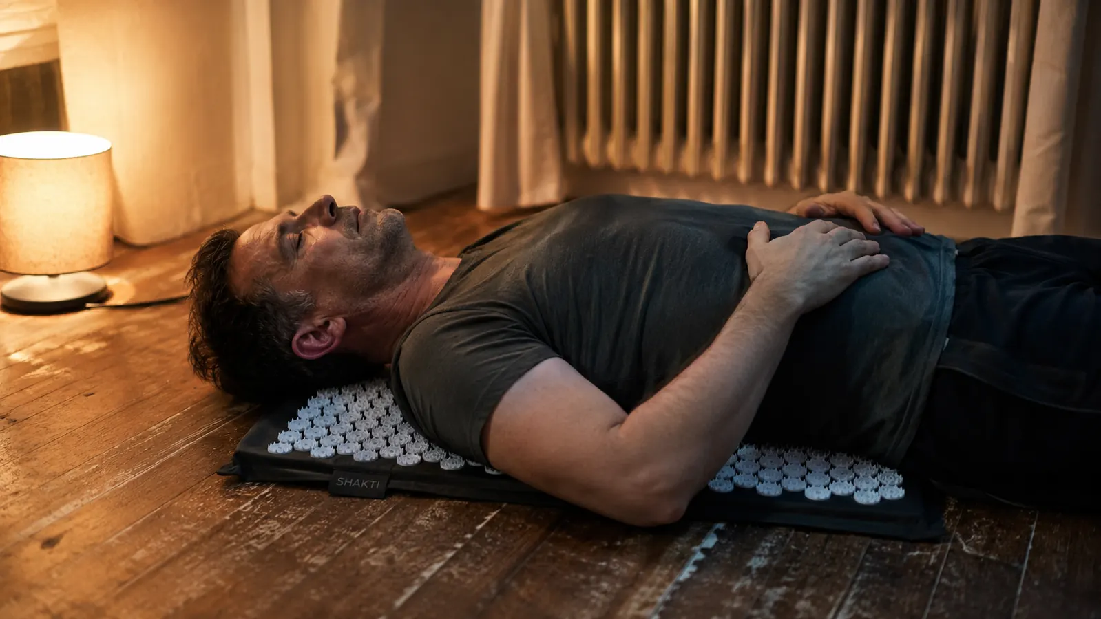 Man lying back on a spiked acupressure mat on his living room floor in the evening, eyes closed, lamp light in the background