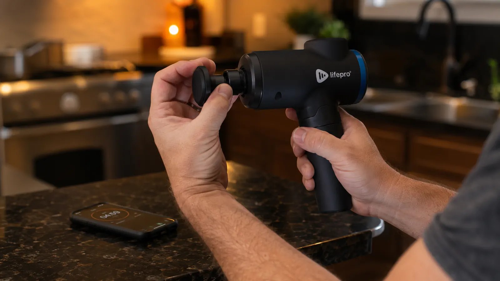 Man switching massage gun attachment heads on a kitchen counter before an evening recovery session