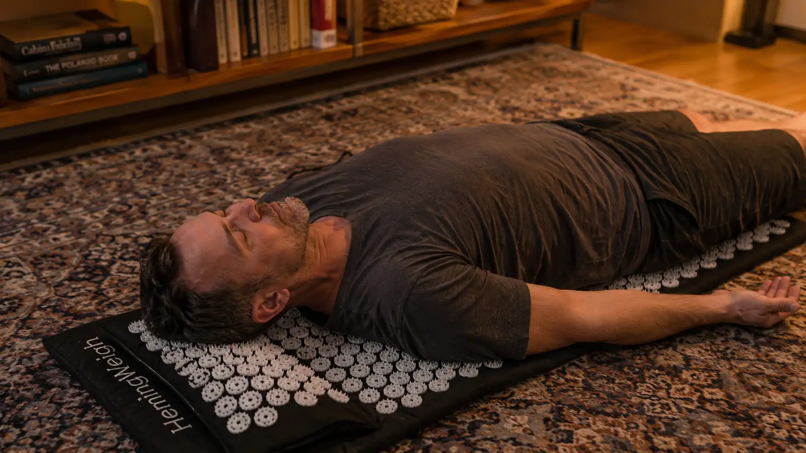 Man in his 40s lying back on an acupressure mat on the living room floor with eyes closed, evening light
