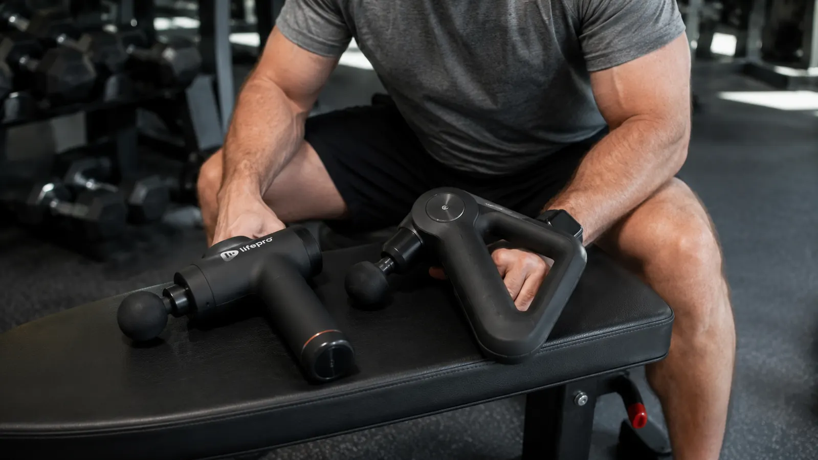 Two percussion massage guns, one in each hand, compared side by side on a gym bench
