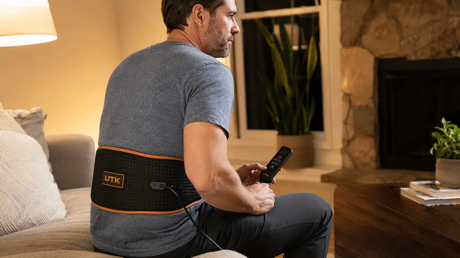Far infrared heating pad wrapped around a man's lower back while he sits on the edge of a couch